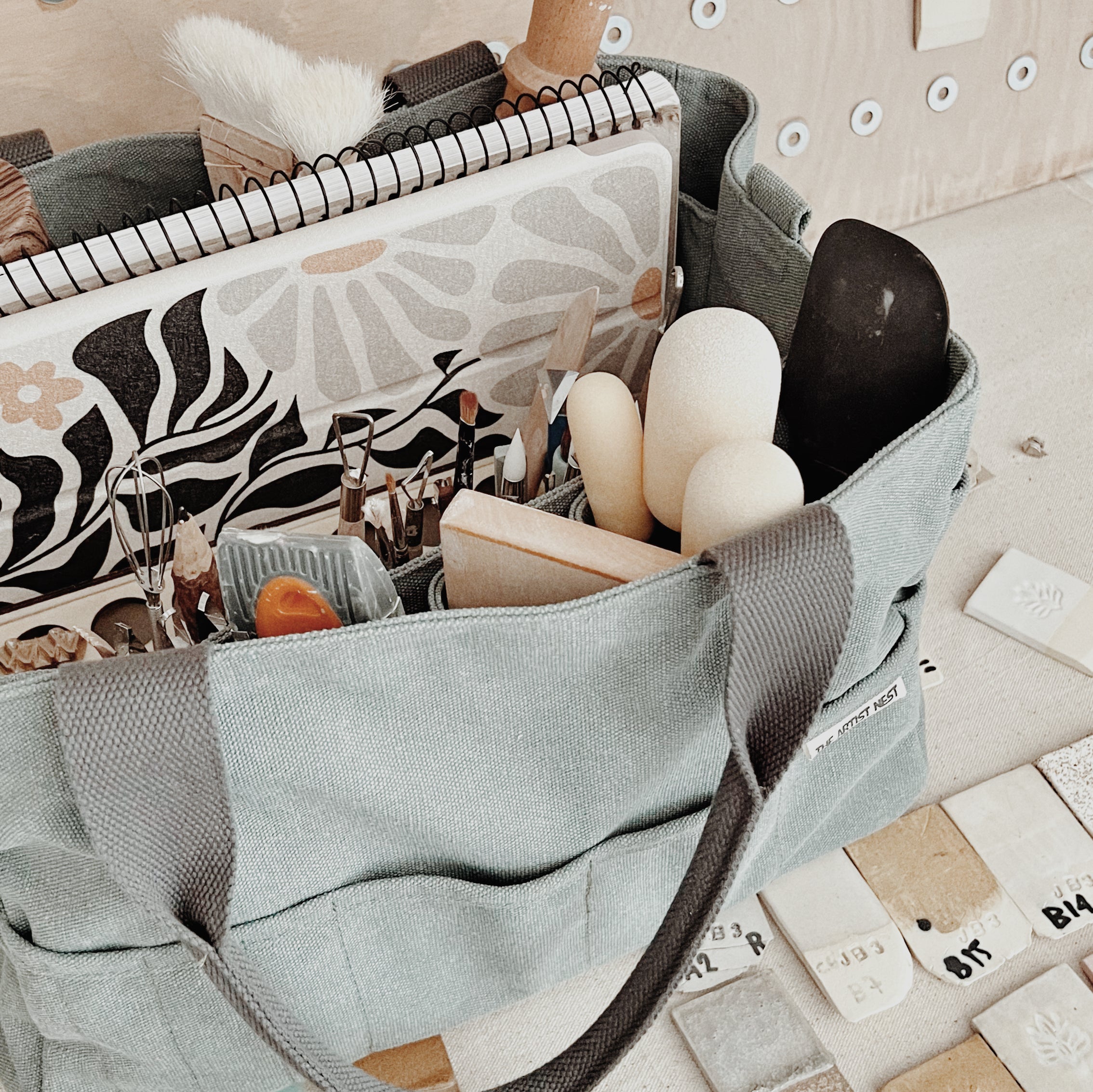 Green Eucalypt pottery tool bag filled with pottery tools and an iPad, shown on a studio table with textiles in a ceramic workspace.