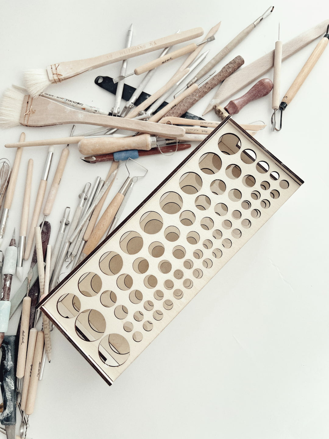 Top view of a wooden pottery tool rack showing custom-sized holes, with pottery tools and pencils scattered on the work table.