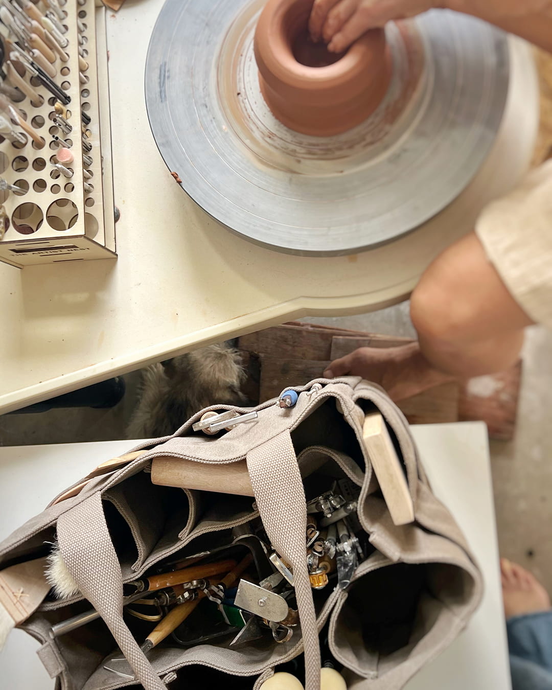 Paperbark Artist Bag top view filled with pottery tools while a potter works at the wheel, ideal pottery tool bag for ceramics classes in Australia.