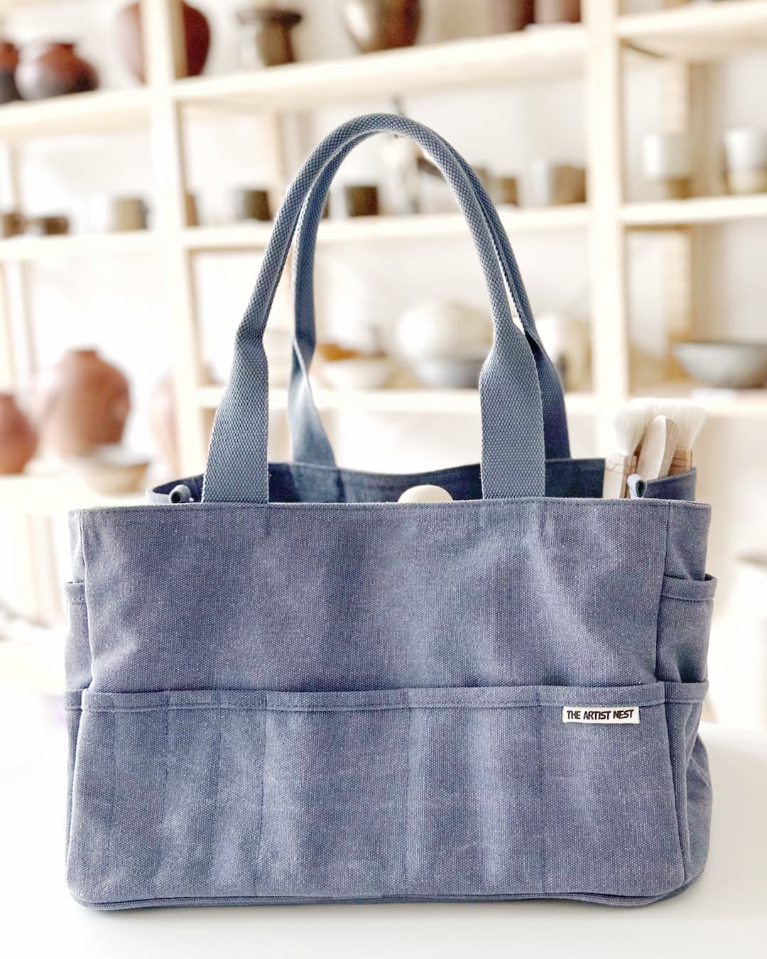 The Pass blue Artist Bag front view styled in a pottery studio with ceramic pots in the background, ideal for ceramic artists needing an organised tool carrier.