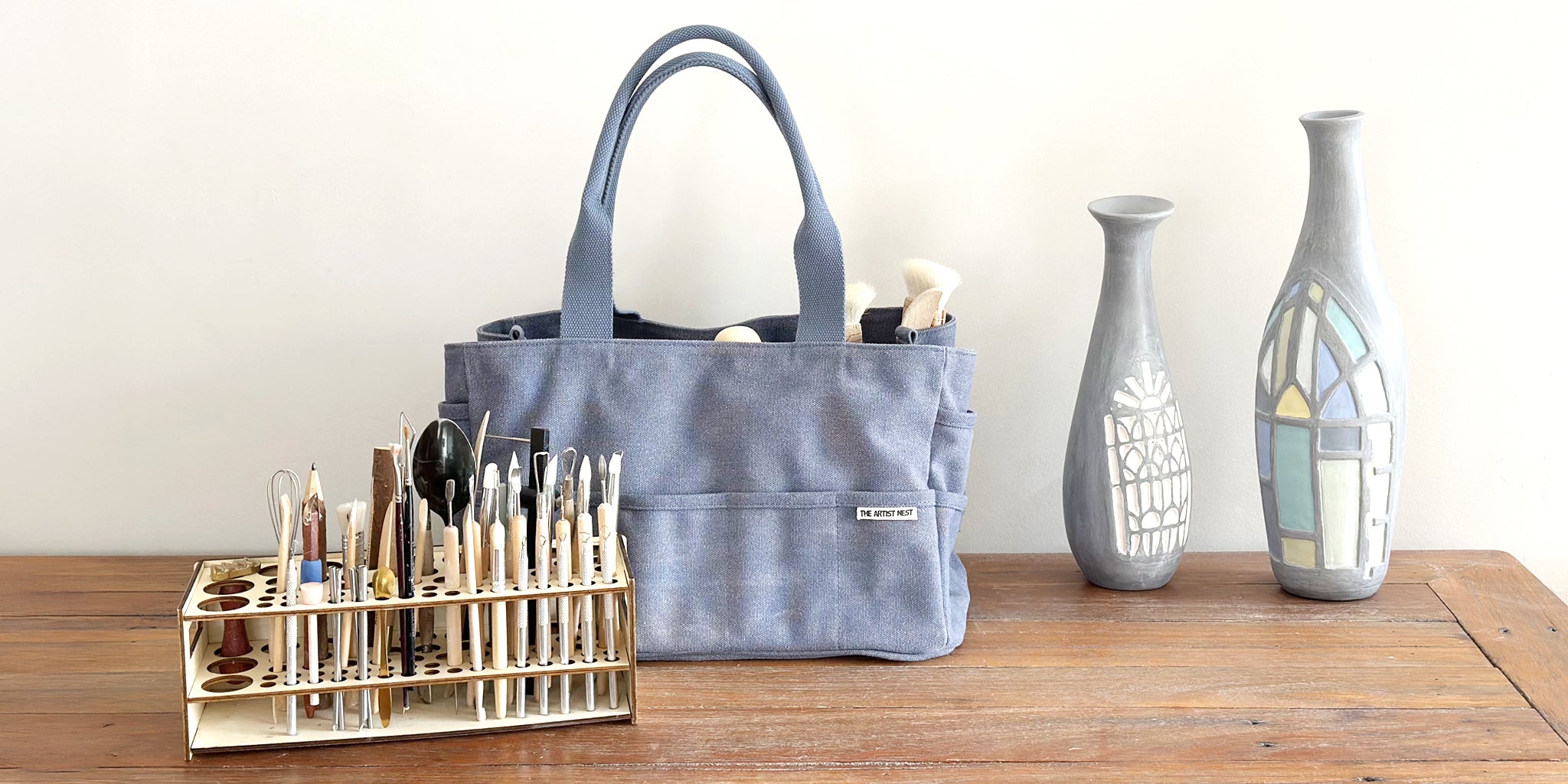The Pass blue artist bag shown with ceramic work, paint brushes, pottery tools, and a wooden tool rack filled with tools.